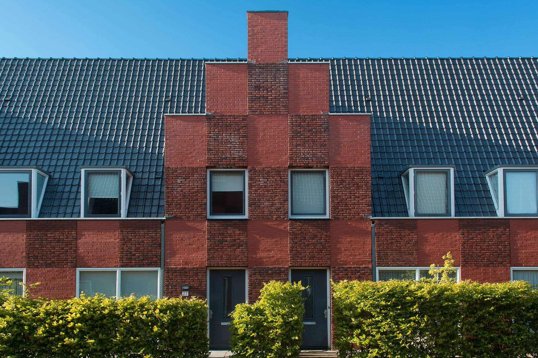 Rijwoningen met een gevel van rode baksteen, donkere dakpannen en hagen voor de deur