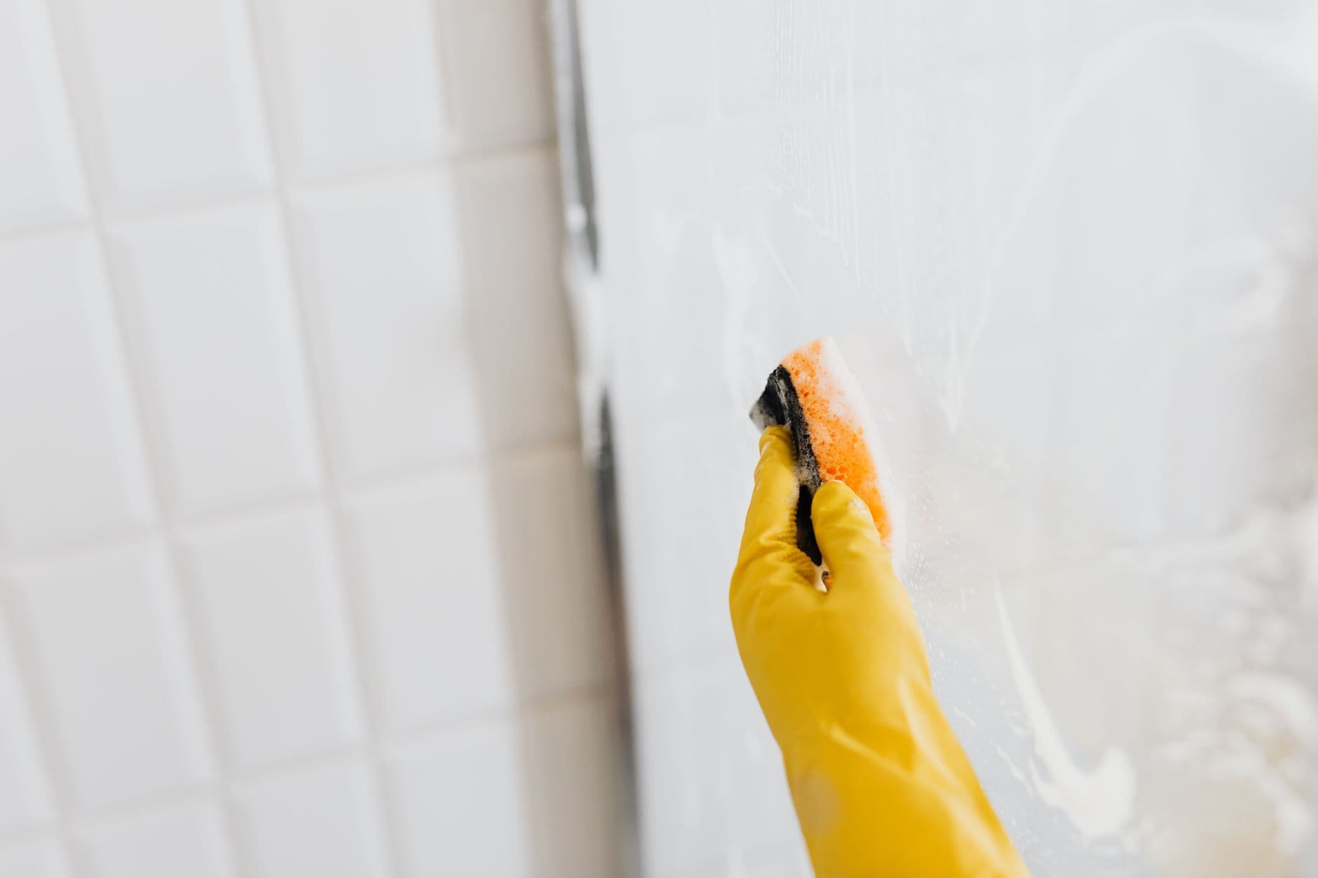 Hand in een gele rubberen handschoen maakt een glazen douchewand schoon met een spons